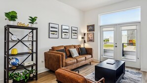 Un salon moderne avec un canapé en cuir marron, une table basse noire et un plancher en bois. On y trouve une étagère noire agrémentée de plantes et d’objets décoratifs.
