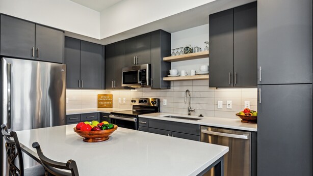Une cuisine moderne avec des meubles de couleur sombre, des appareils en acier inoxydable et un îlot central agrémenté d’un panier de fruits.