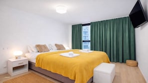 Ein modernes Hotelzimmer mit Bett, Nachttisch, Flachbildfernseher an der Wand und Fenster mit grünen Vorhängen.