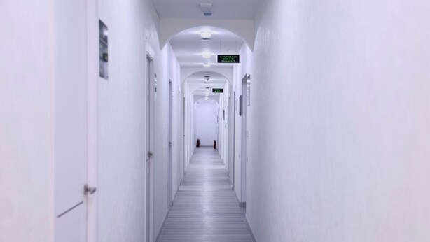 Ένας μακρύς, στενός διάδρομος με λευκούς τοίχους και πόρτες και στις δύο πλευρές, που οδηγεί σε μια απομακρυσμένη έξοδο.