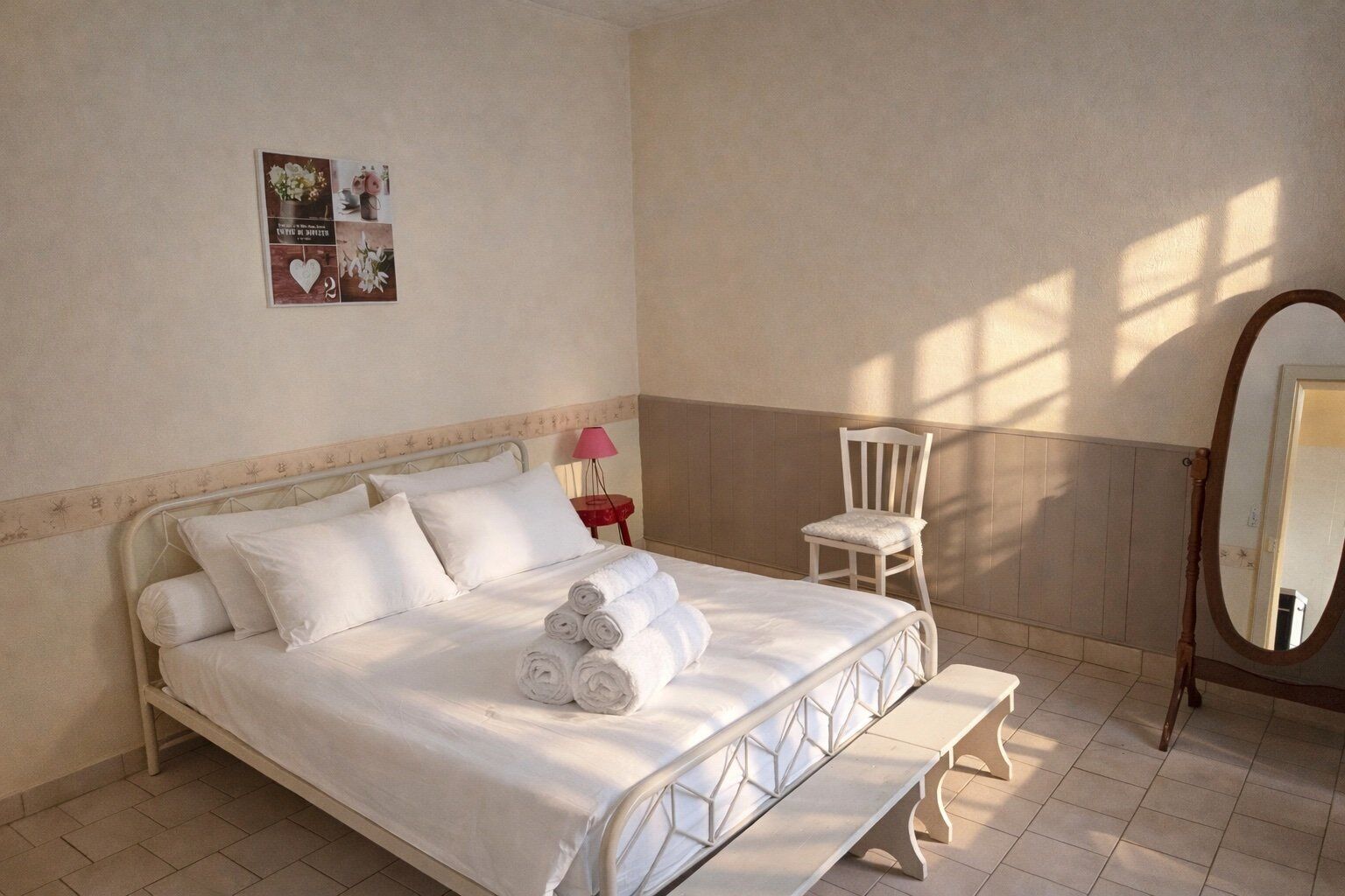 A neatly made bed with rolled towels, a small bedside table with a lamp, a chair, and a mirror.