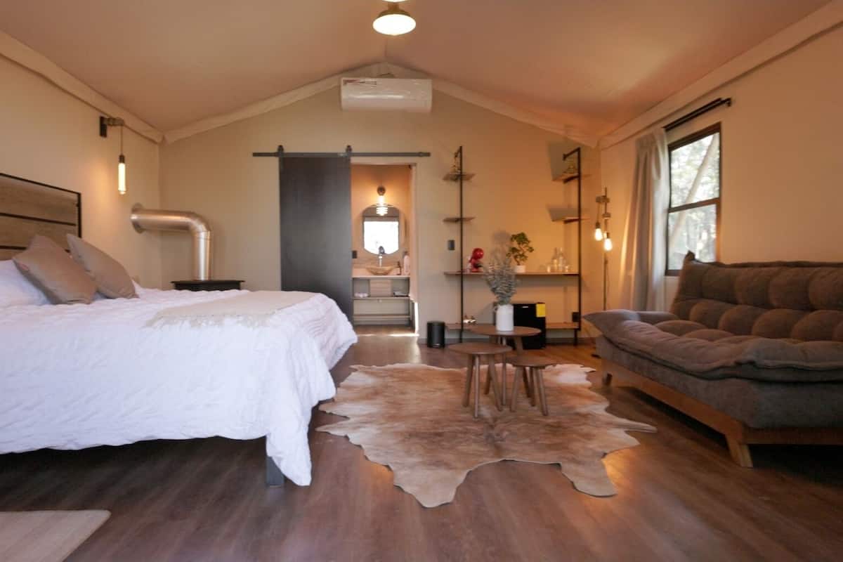 A bedroom with a large bed, a sofa, a dining table, and a kitchen area in the background.