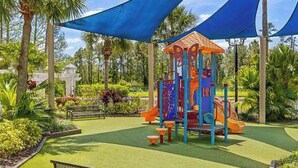 Un parque infantil con un colorido arenero y un tobogán, rodeado de follaje y palmeras.