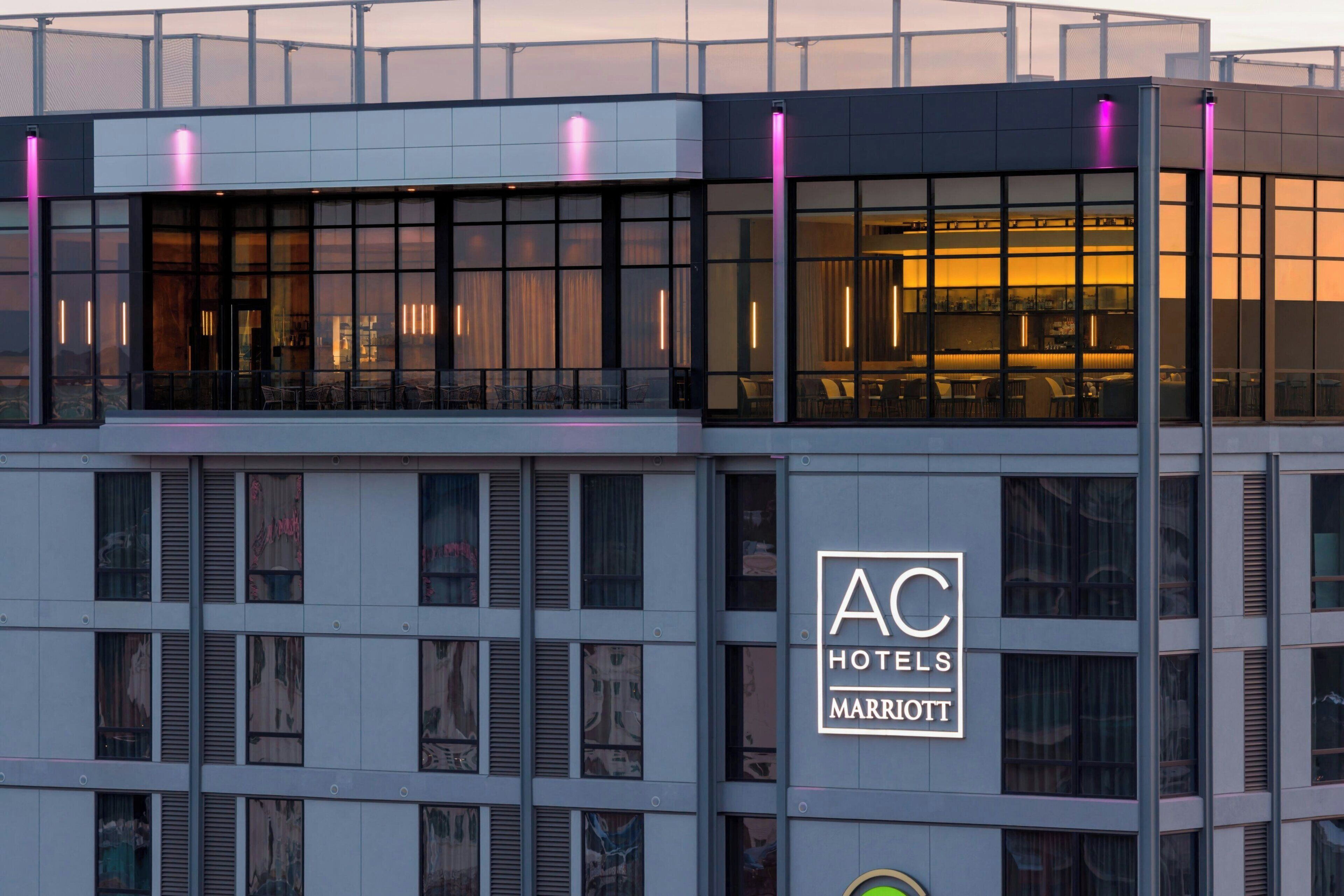 A modern hotel building with a prominent 'AC HOTELS MARRIOTT' sign, large windows, and a balcony area.