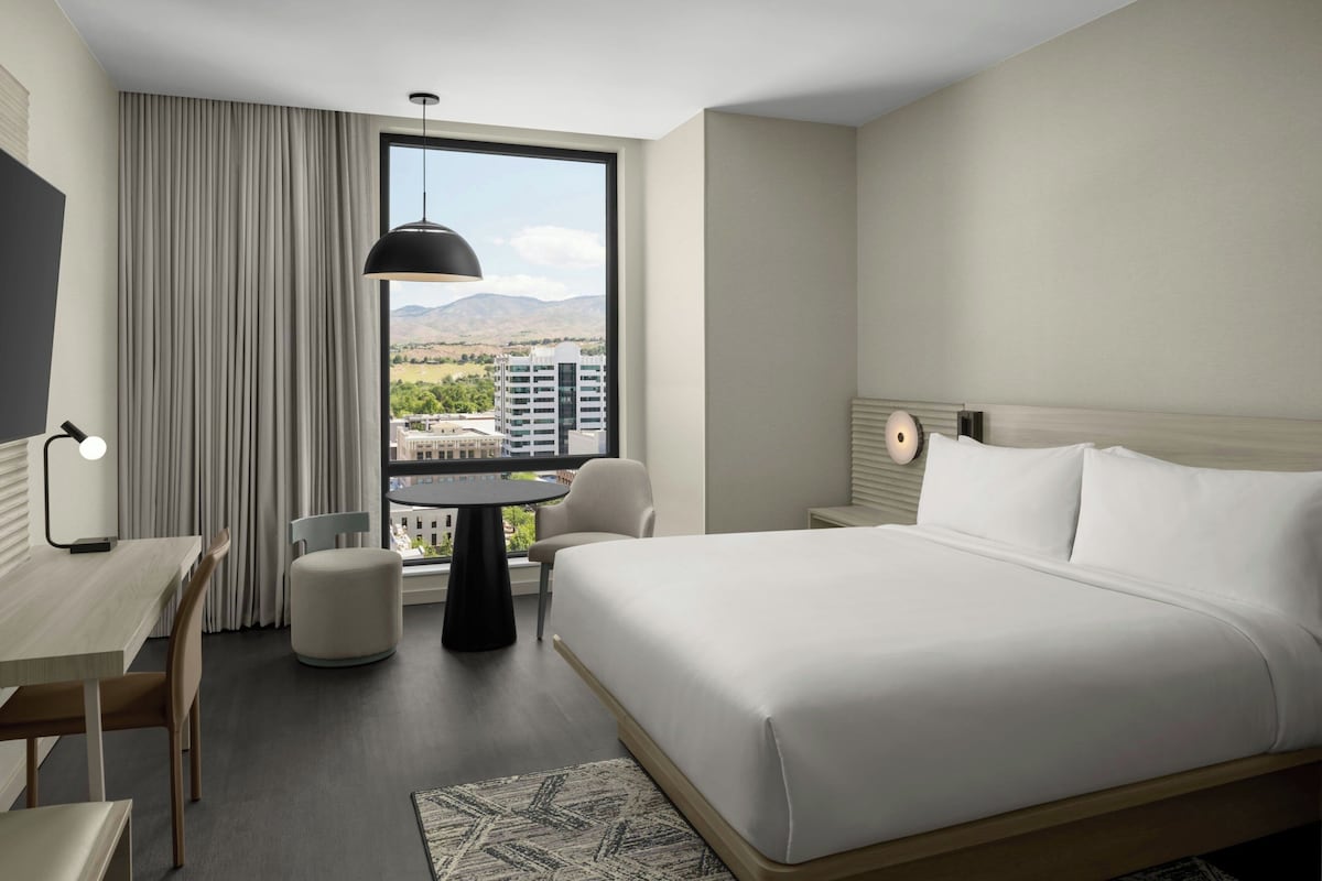 Une chambre d’hôtel avec un lit, un bureau, une chaise, une table et une vue sur le paysage urbain, visible à travers une grande fenêtre.