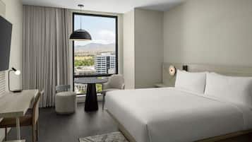 A hotel room with a bed, a desk, a chair, a table, and a view of a cityscape through a large window.