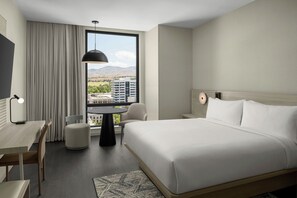 Ein Hotelzimmer mit einem Bett, einem Schreibtisch, einem Sessel, einem Tisch und Blick auf eine Stadtlandschaft durch ein großes Fenster.
