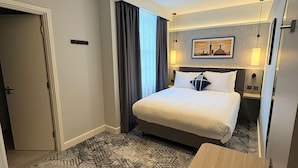 Une chambre d’hôtel comprenant un lit, un bureau, une chaise, une fenêtre avec des rideaux, une lampe fixée au mur et un tableau.