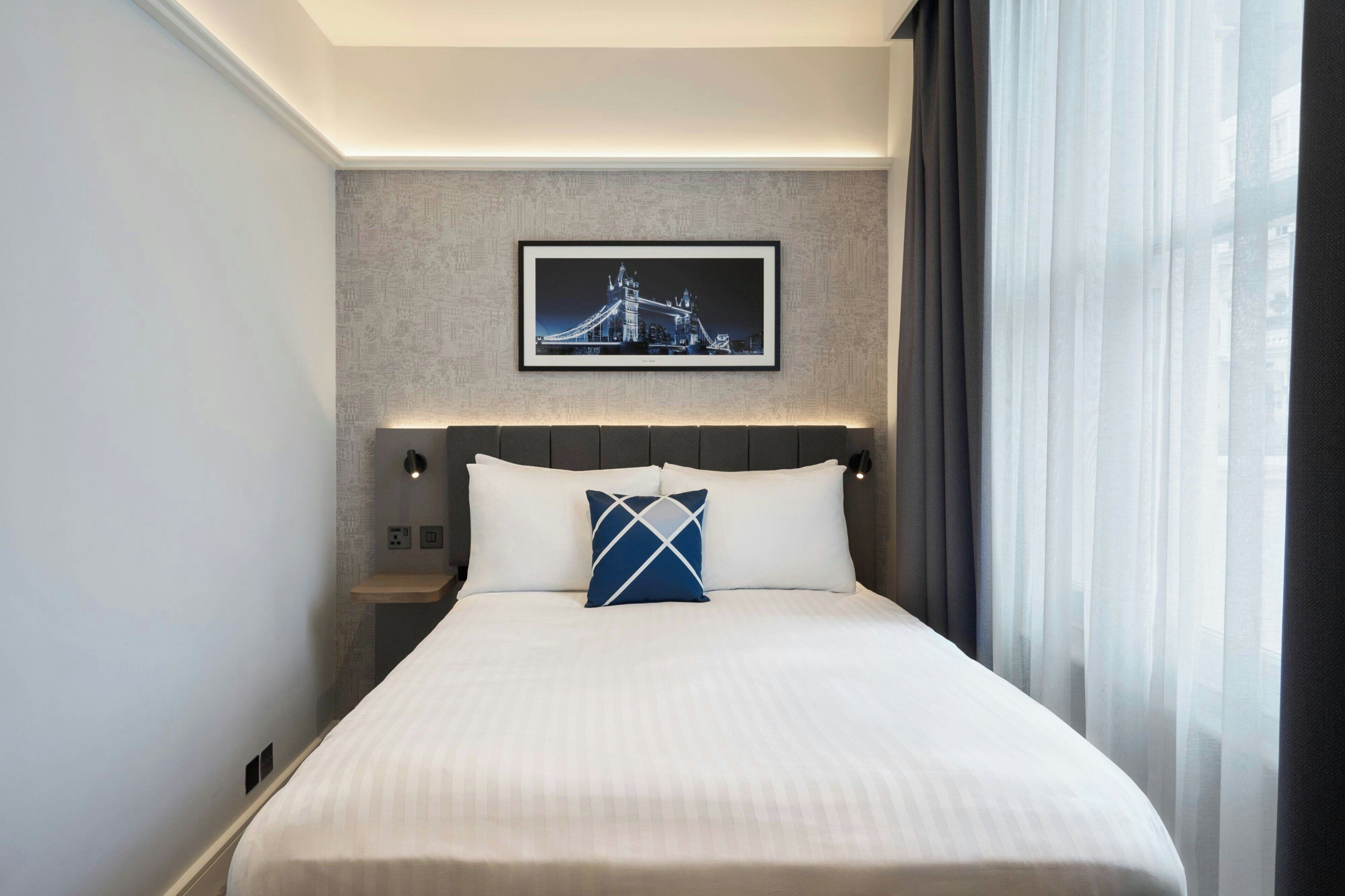 Een hotelkamer met een bed, een nachtkastje, een ingelijst schilderij aan de muur en een raam met gordijnen.