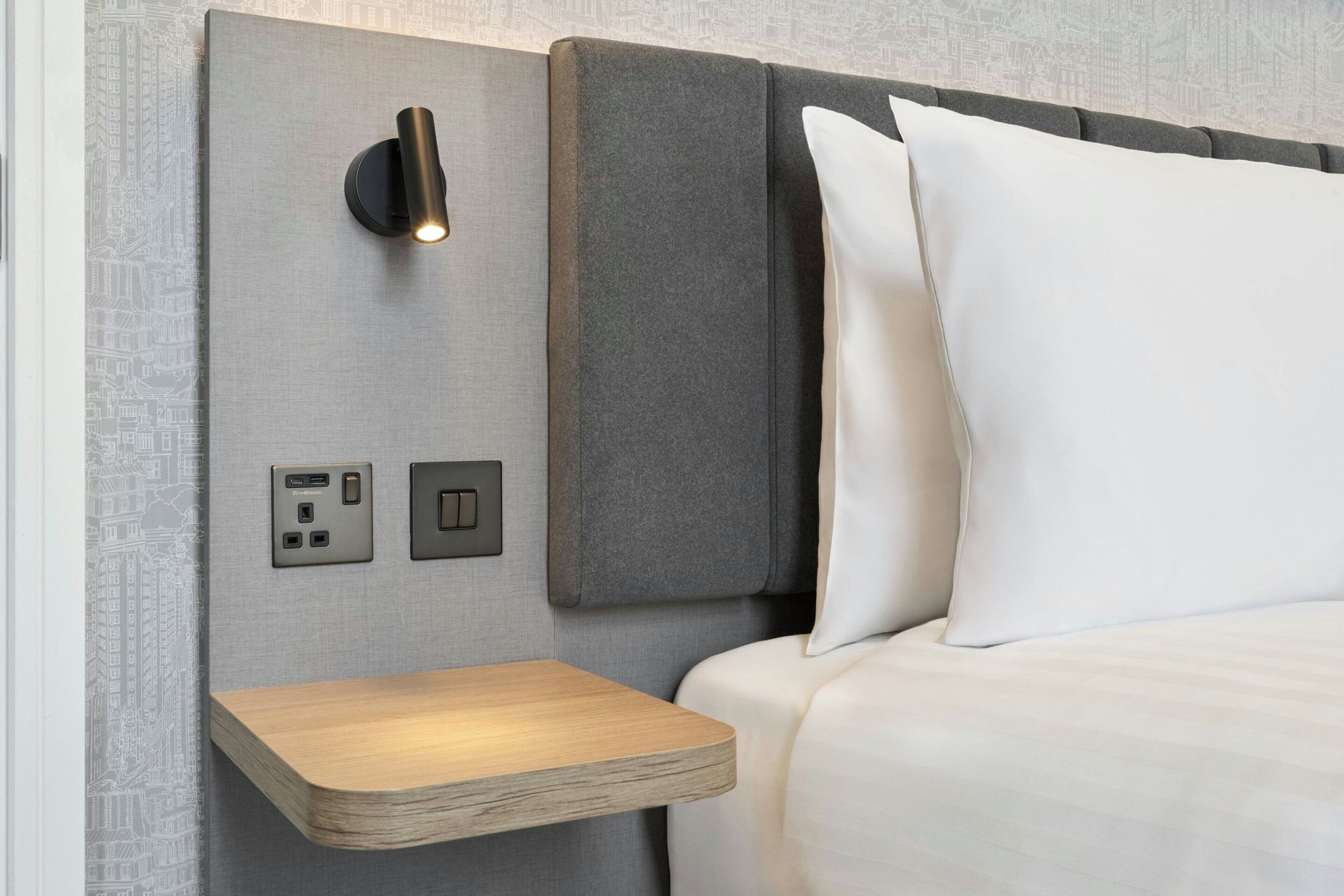 A neatly made bed with white linens, a bedside table with a light, and a wall-mounted switch.