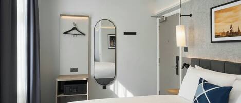 A hotel room with a bed, a bedside table, a mirror, a hanging coat rack, and a wall-mounted picture.