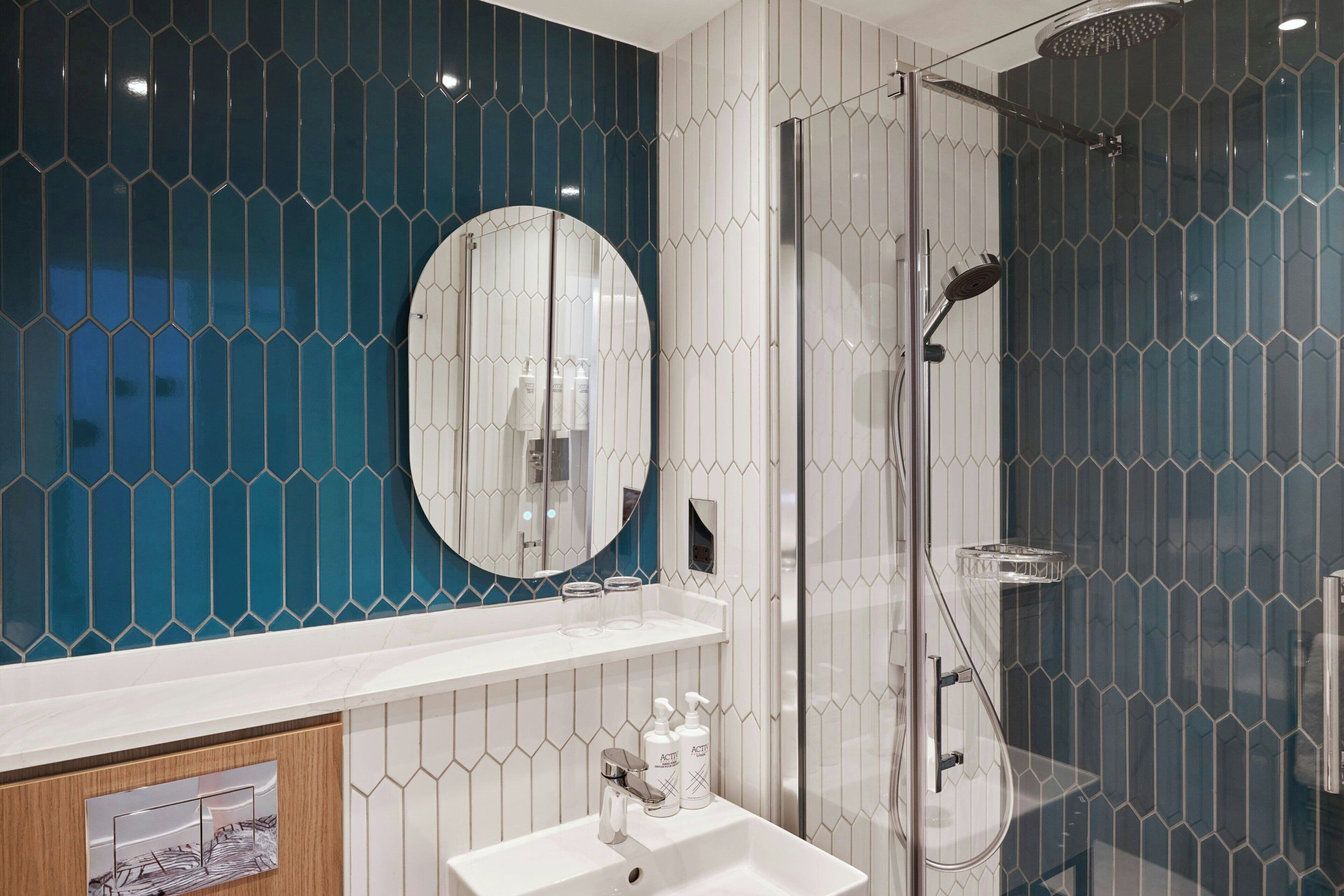 A modern bathroom with a glass-enclosed shower, a round mirror, and a white sink with a wooden cabinet below.