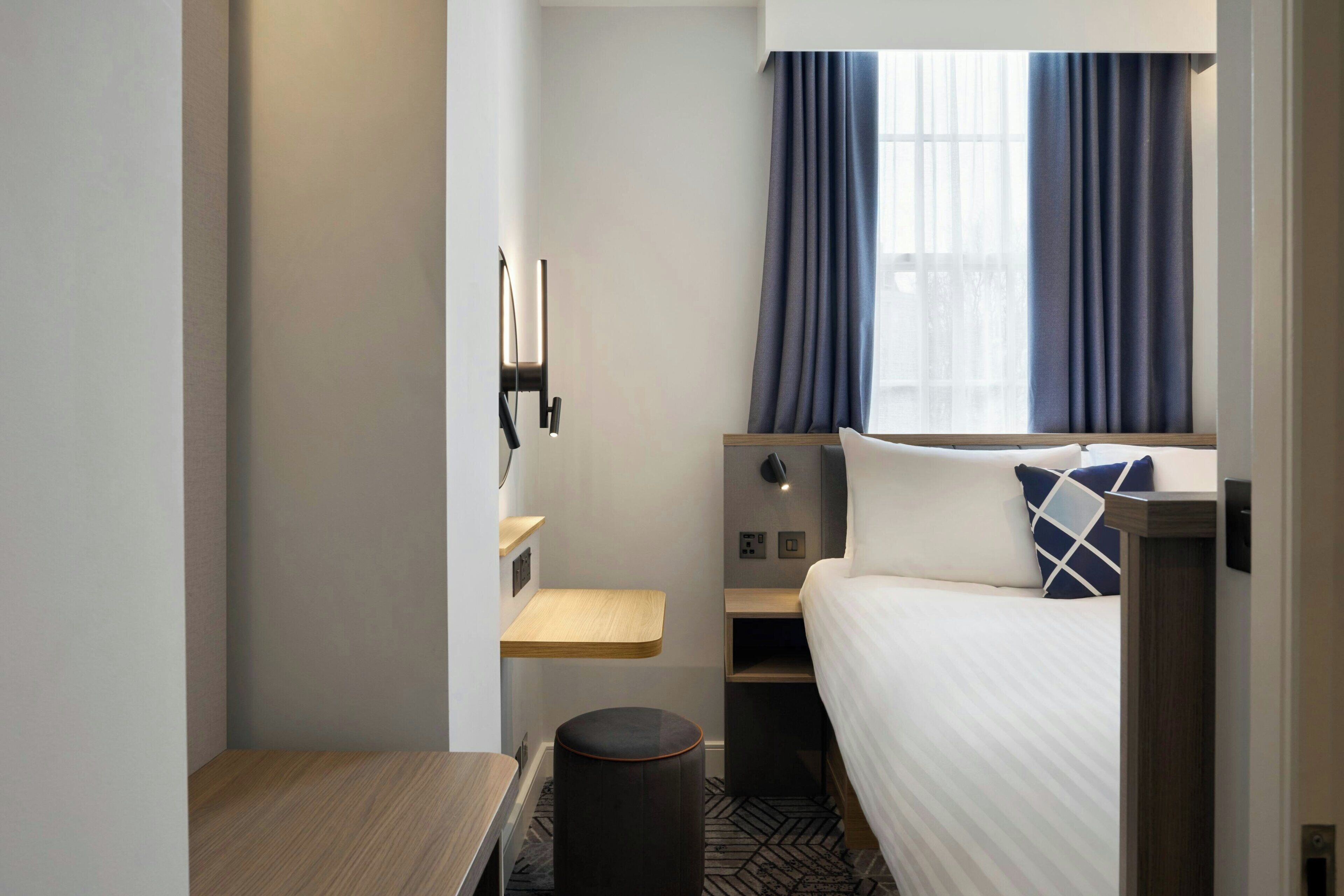 A compact hotel room with a bed, a small table, a shelf, and a window with curtains.