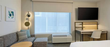 A modern hotel room with a sofa, a small round table, a desk with a chair, a flat-screen TV, and a large window with blinds.