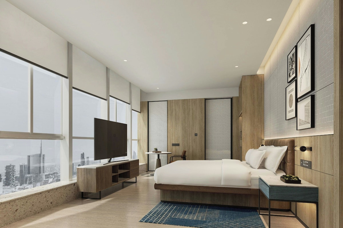 A modern hotel room with a large bed, a TV, a desk, and a city view.