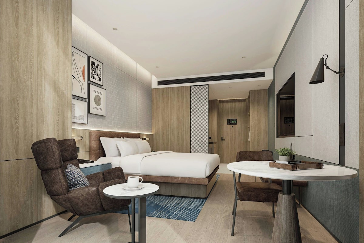 A modern hotel room with a bed, two armchairs, a small round table, and a TV.
