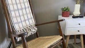 Un fauteuil à bascule en bois avec une couverture à carreaux, une petite table d’appoint blanche avec une lampe, et une petite plante en pot.