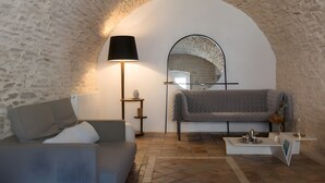 Une pièce chaleureuse avec un mur en pierre, un canapé gris, une petite table avec une lampe et un miroir.