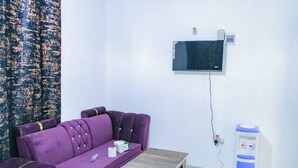 A room with a purple sofa, a small wooden table, a TV mounted on the wall, and a water dispenser. - Yomoke Hotel Miritini (Mombasa)