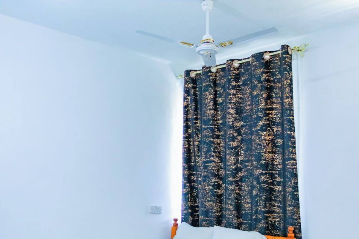 A single bed with a wooden frame and white bedding in a room with white walls, a ceiling fan, and a window with dark patterned curtains.