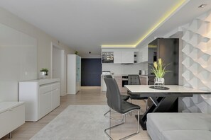 A modern kitchen with white cabinets, a dining area with a white table and black chairs, and a living area with a grey sofa. - Szafarnia 2 by Grand Apartments (Gdańsk)