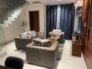 A modern living room with a staircase, grey sofas with patterned cushions, a wooden coffee table, and a fan. - Yaba PentHouse Apt 2B2B with Security and Lift and Generator (Lagos)