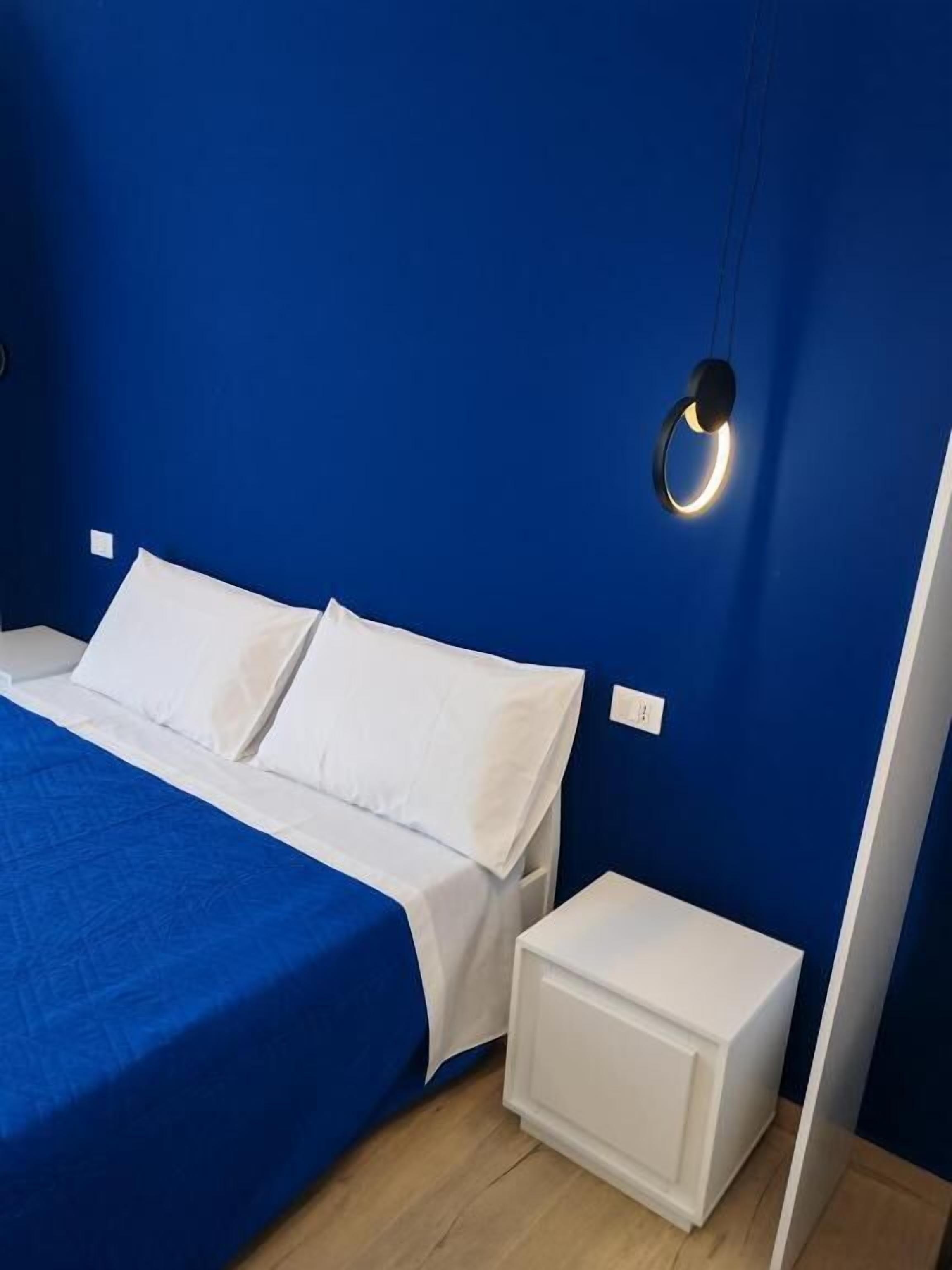 A bedroom with a blue wall, a bed with white bedding, a bedside table, and a circular light fixture.
