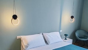 A neatly made bed with a teal bedspread and white pillows, two bedside tables with lamps, and a chair in a room with light blue walls and wooden flooring. - Mare Suite (Palermo)
