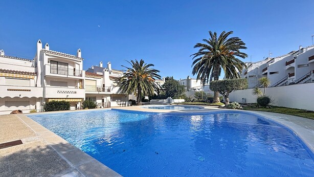 A swimming pool with a clear blue water surface, surrounded by white buildings with balconies and palm trees. - JARDINS I BX 27 Roses / Roses - INTERimmo (Rosas / Roses)