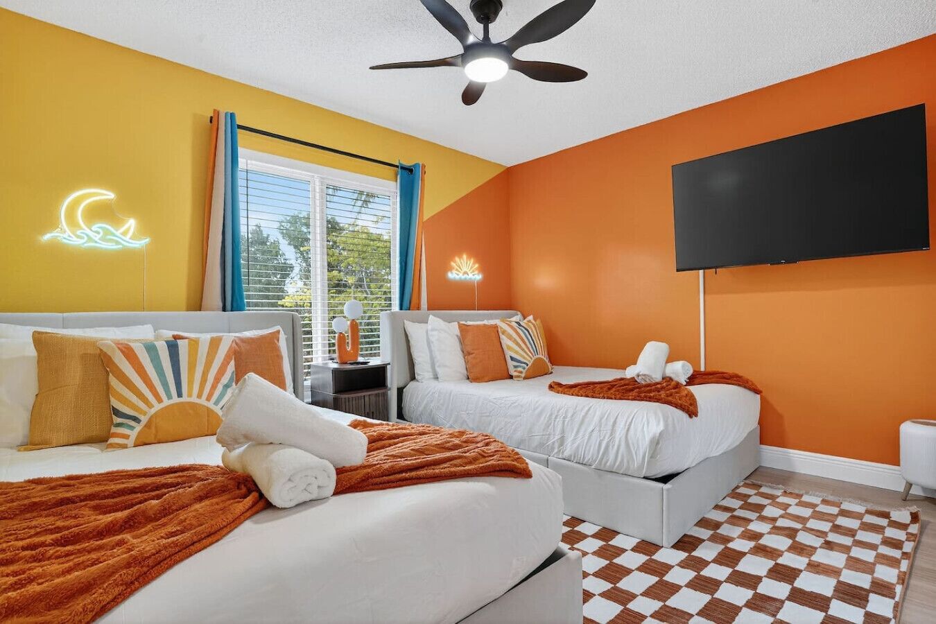 Quarto de hotel com duas camas, um ventilador de teto, televisão e piso xadrez.