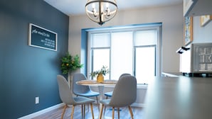 A dining area with a round table, four chairs, a chandelier, a window with blinds, and a wall-mounted sign.
