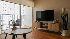Ein modernes Wohnzimmer mit großem Fernseher, Holzmöbeln, einem Balkon mit Blick auf Gebäude und einem Blumentopf.
