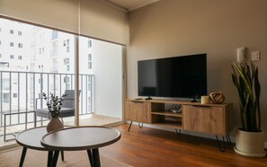 Uma sala de estar moderna com uma televisão grande, móveis de madeira, uma varanda com vista para edifícios e um vaso de planta.