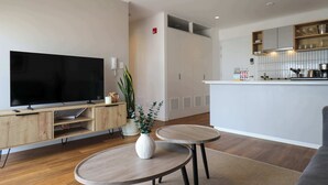 Ein modernes Wohnzimmer mit Flachbildfernseher, Holzmöbeln, einer kleinen Essecke und einer Küche mit weißen Schränken und Arbeitsplatte.