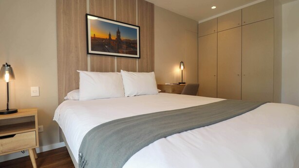 Ein Hotelzimmer mit einem großen Bett, zwei Nachttischlampen, einem Nachttisch, einem Stadtbild im Bilderrahmen und einem Kleiderschrank.