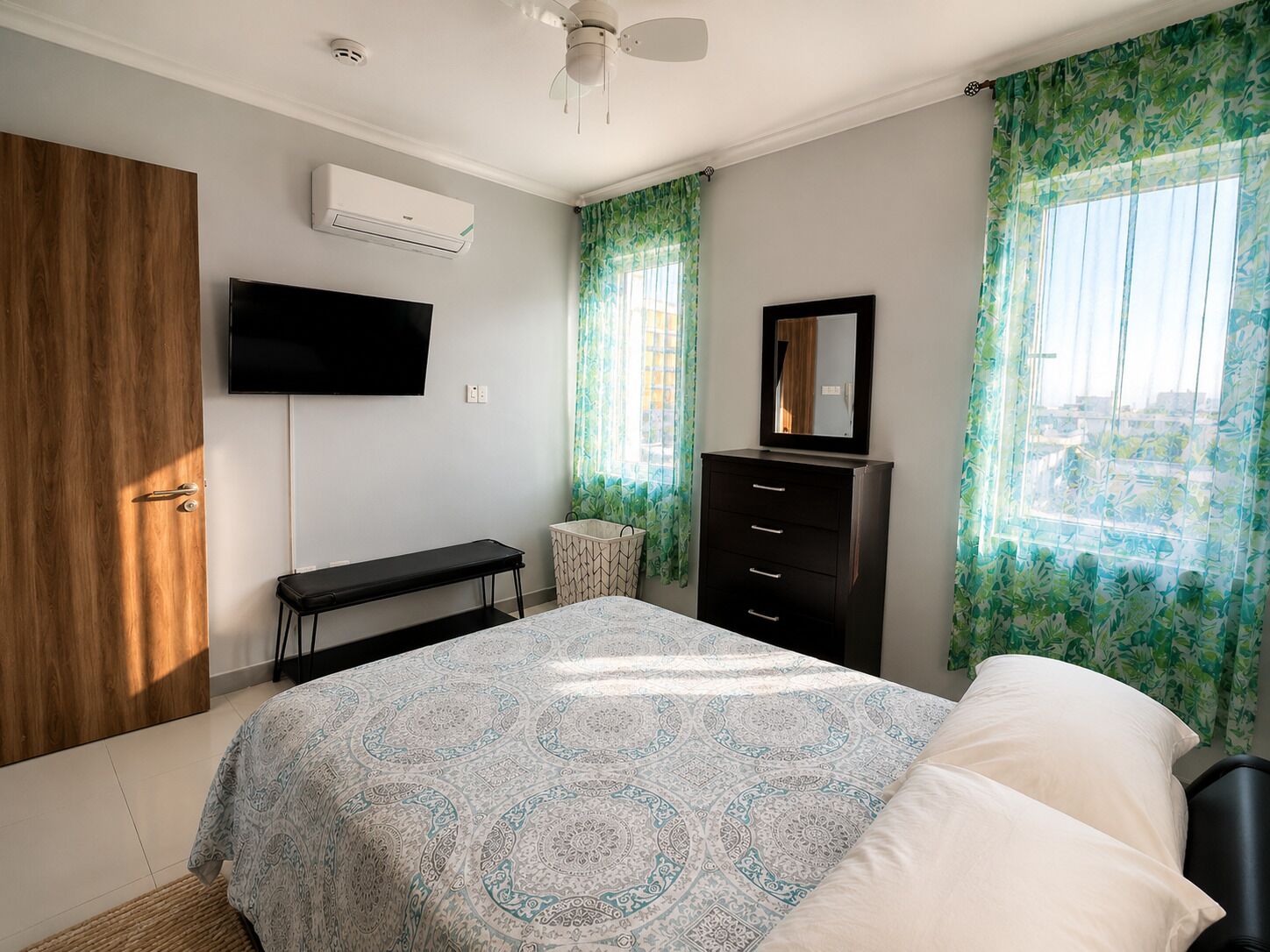 Une chambre à coucher avec un lit, une télévision, une commode, un banc et une fenêtre avec des rideaux.