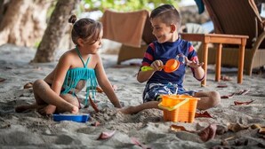 Two children playing with sand on a beach, with beach chairs and a table in the background. - Arenas del Mar Beachfront & Rainforest Resort (Quepos)