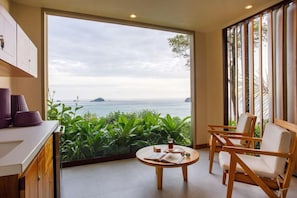 A room with a large window offering a view of the sea and greenery. There is a round wooden table with a book and a glass. - Arenas del Mar Beachfront & Rainforest Resort (Quepos)