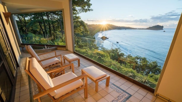 A balcony with wooden furniture, a tiled floor, and a view of the ocean and forest. - Arenas del Mar Beachfront & Rainforest Resort (Quepos)