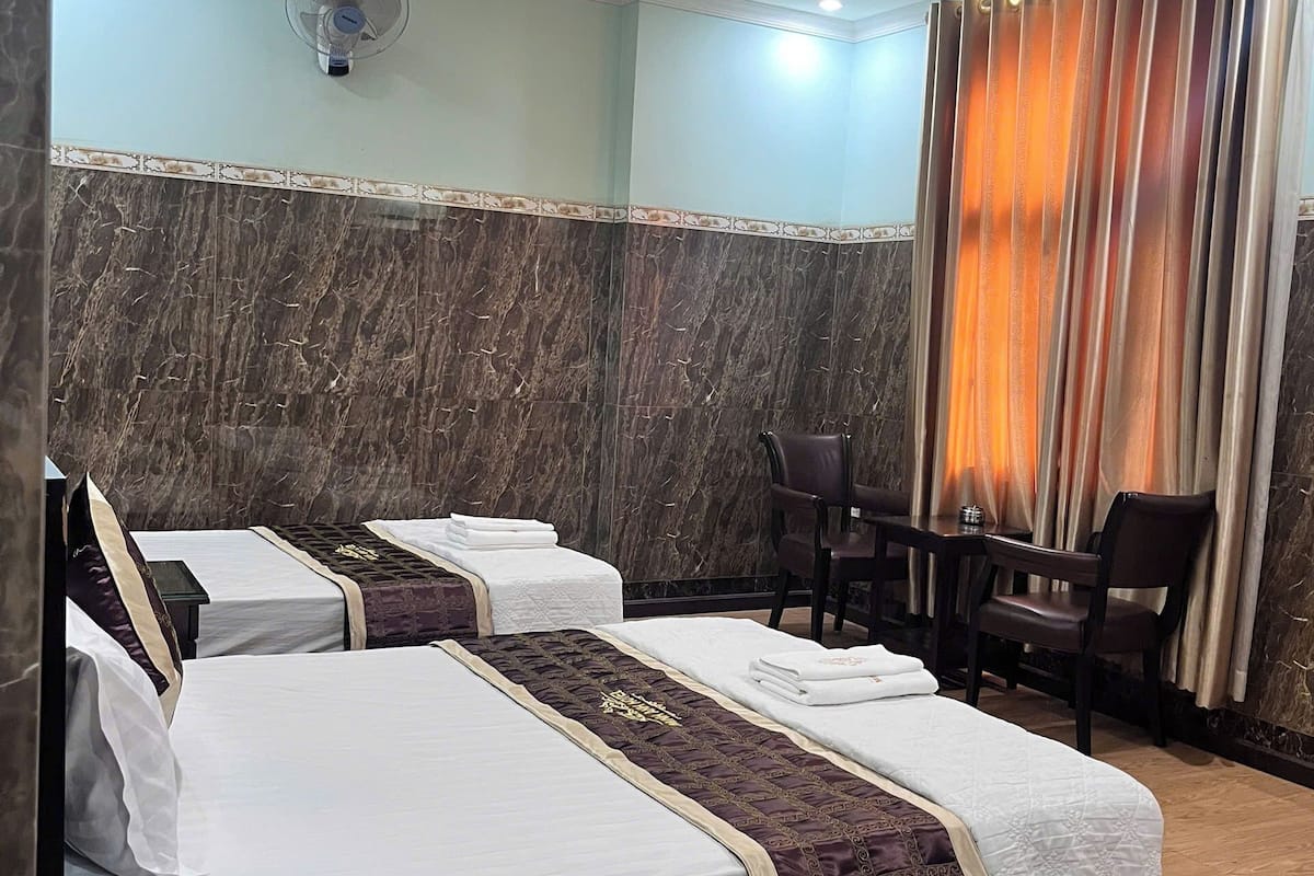 Ein Hotelzimmer mit drei Betten, einem Schreibtisch, Stühlen, einem Ventilator und Blick nach draußen.