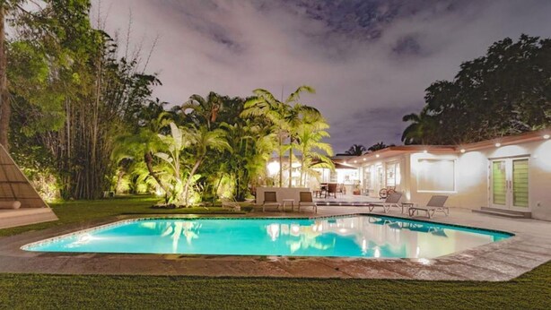 A swimming pool with a grassy area and palm trees, surrounded by a building with outdoor seating and a covered patio. - Villa Oahu Basketball Fire Pit Mini Golf (North Miami Beach)
