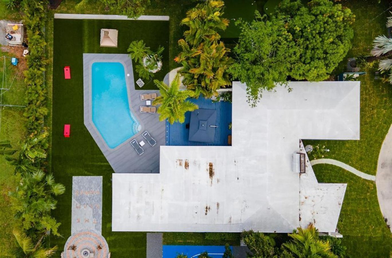 Vista aérea de una propiedad con piscina, sillas de descanso y un edificio grande de color blanco con techo azul.