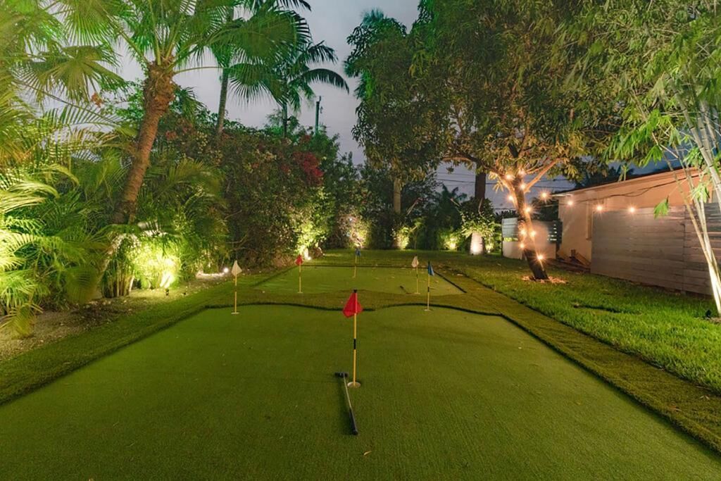 Ein gut beleuchteter Minigolfplatz im Abendlicht mit einer roten Fahne in der Mitte, umgeben von üppigem Grün und Bäumen.