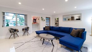 A modern living room with a blue sectional sofa, two armchairs, a coffee table, and a large window providing natural light. - Villa Oahu Basketball Fire Pit Mini Golf (North Miami Beach)