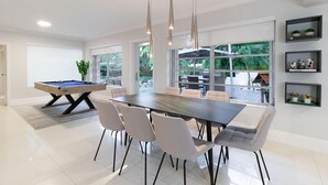 A modern dining area with a pool table, a long dining table with chairs, and a view of the outdoors through large windows. - Villa Oahu Basketball Fire Pit Mini Golf (North Miami Beach)