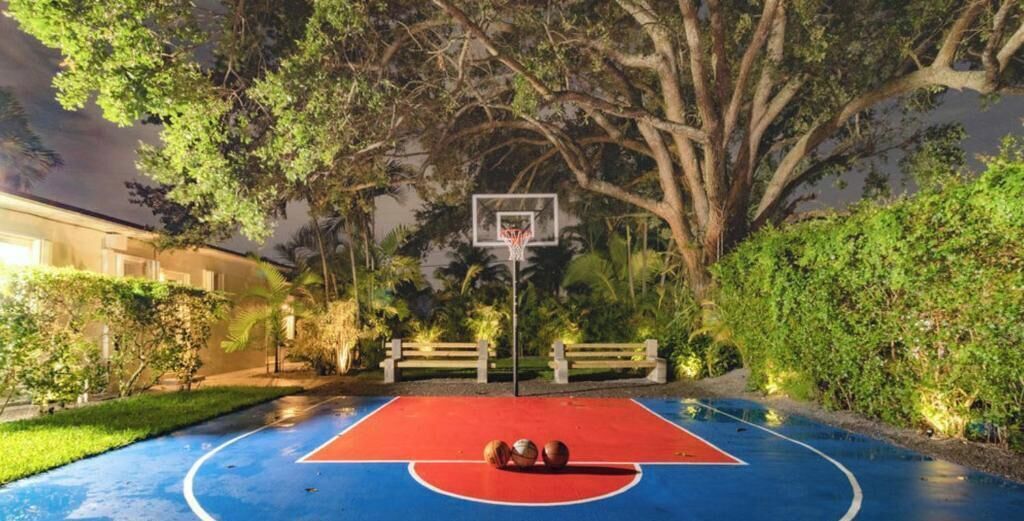 Quadra de basquete ao ar livre com cesta, cercada por árvores e um banco.