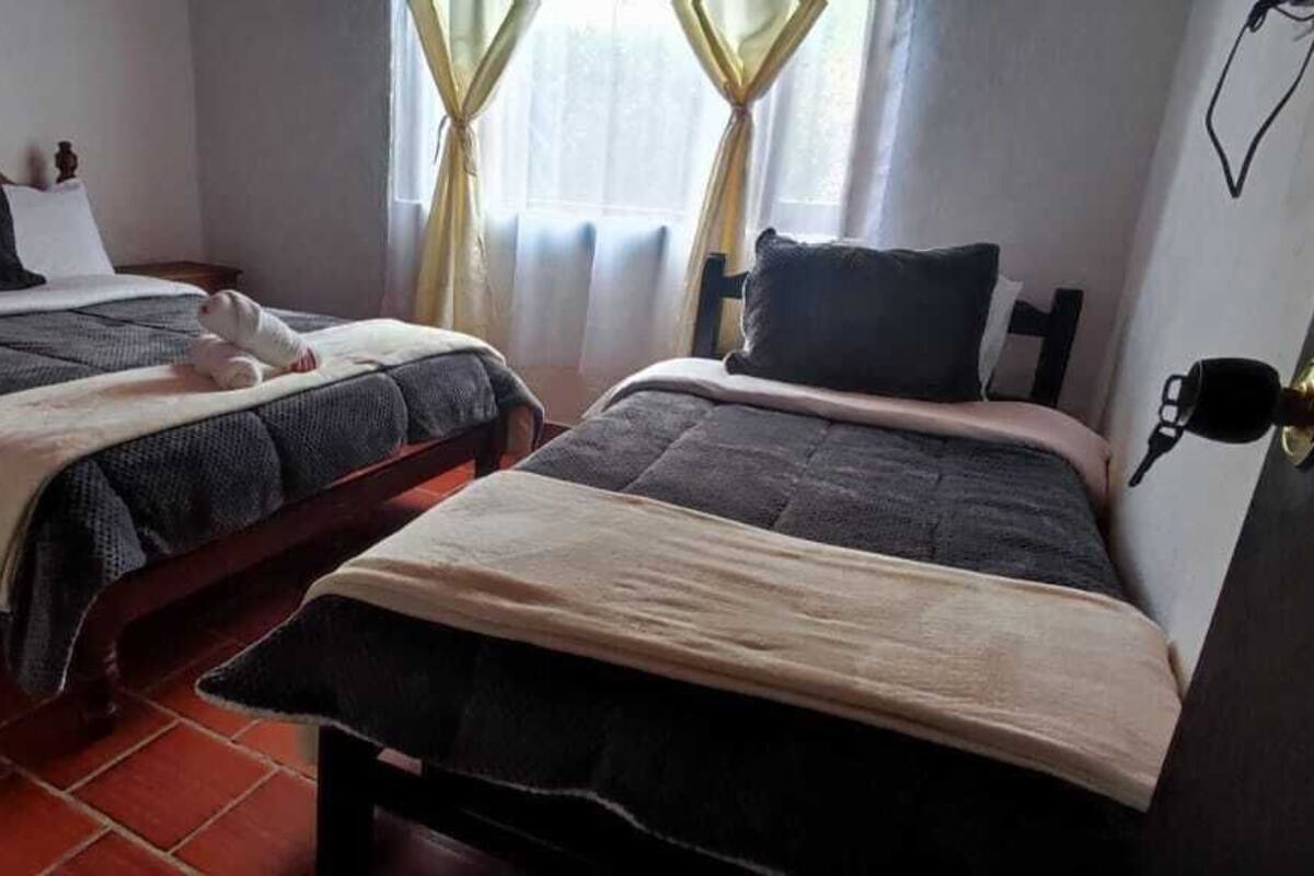 A room with two beds, each with a different style of headboard. There is a window with curtains, a door with a handle, and a wall-mounted light fixture.
