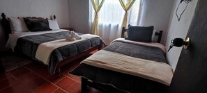 A room with two beds, each with a different style of headboard. There is a window with curtains, a door with a handle, and a wall-mounted light fixture. - Cabaña Lewana Campestre (Villa de Leyva)