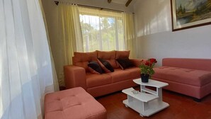 A living room with a sofa, ottoman, and coffee table, a painting on the wall, and a window with curtains. - Cabaña Lewana Campestre (Villa de Leyva)