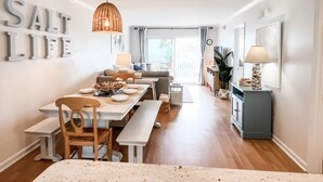 A bright, open-plan living and dining area with a large window, a dining table set for four, and a cozy seating area with a sofa and chairs. - Modern Beachfront Condo w/pool Carolina Beach (Carolina Beach)